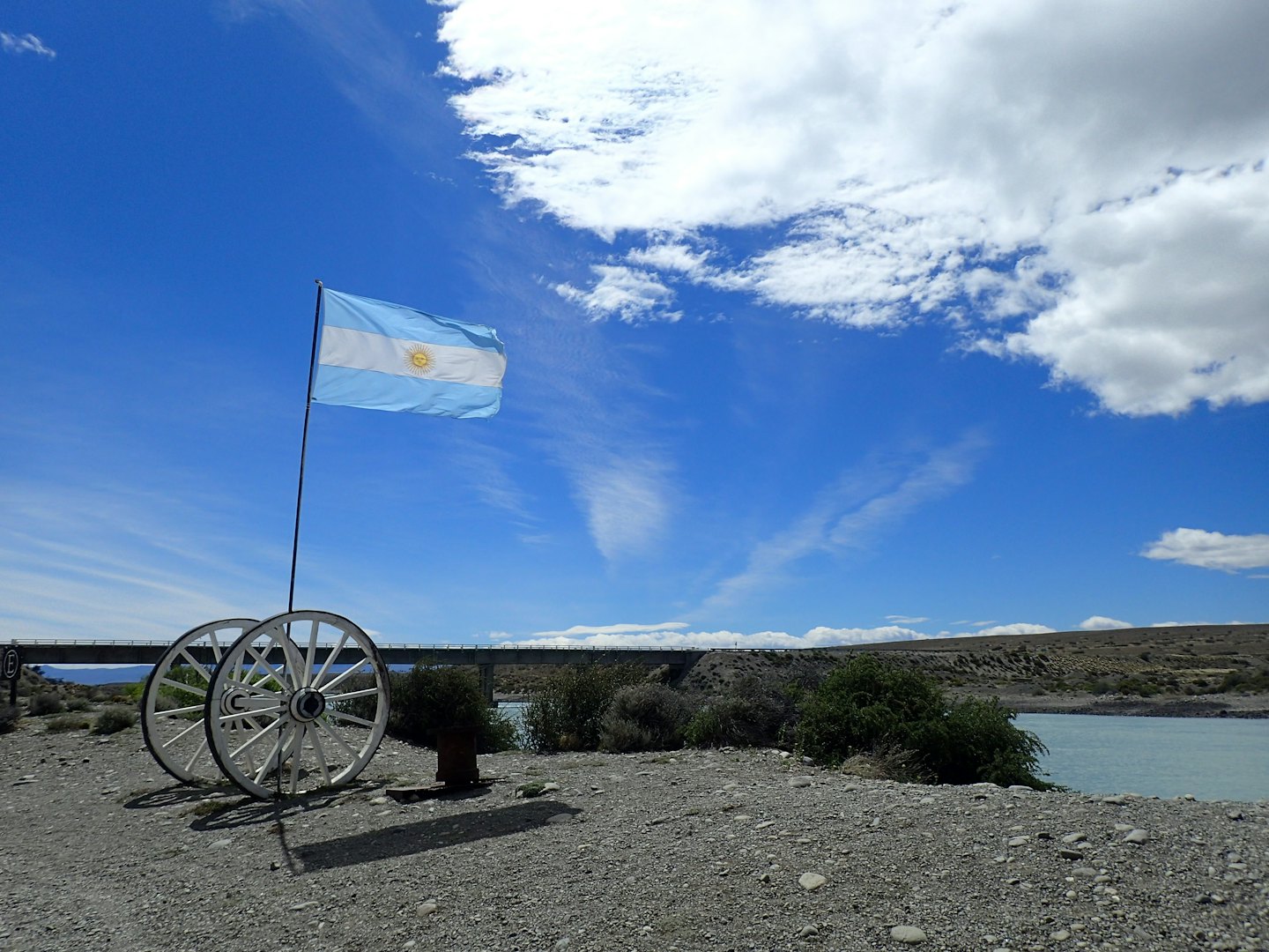 Argentinian flag waving between white wagon wheels near a river.